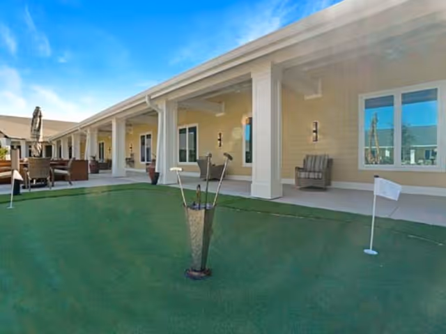 Covered patio with seating and a small putting green in front of a row of ground-floor residential units.