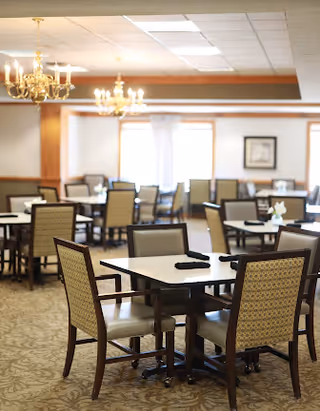 Bright dining room with multiple square tables and upholstered chairs, chandeliers, and simple place settings.