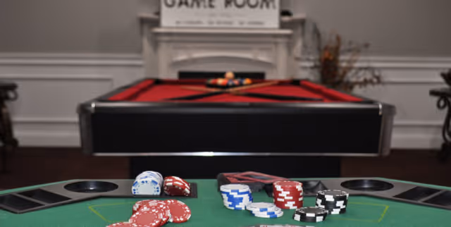 A game room featuring a poker table with poker chips and card holders in the foreground, and a red felt pool table with billiard balls and cues in the background. The room has white paneled walls and a sign above the fireplace that reads 'GAME ROOM'.