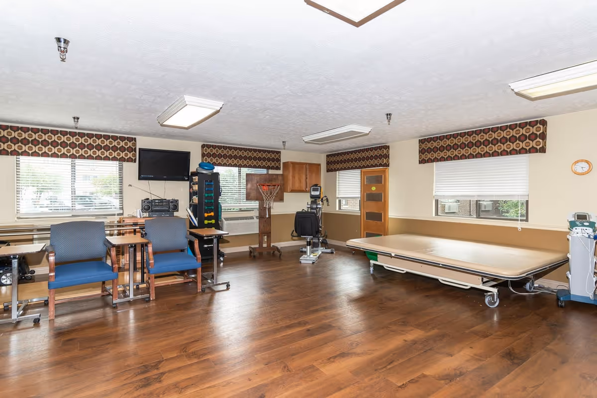 A spacious room with wooden flooring featuring two blue chairs with wooden arms, small tables, a flat-screen TV mounted on the wall, exercise equipment including a stationary bike and a basketball hoop, and a large medical examination or therapy table. The room has three windows with patterned valances and white blinds, beige walls with a brown chair rail, and ceiling lights.