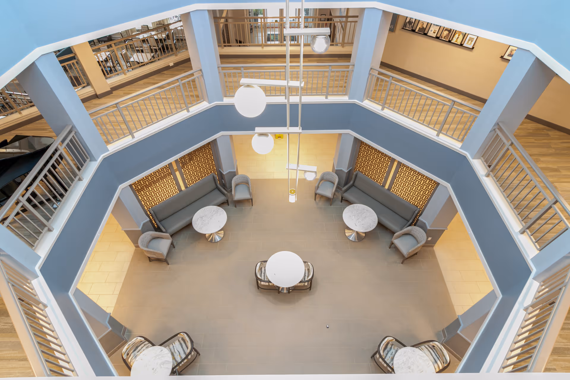 View from above of a spacious, octagonal indoor seating area with blue walls and railings on the upper level. The seating area includes sofas, armchairs, and round tables with marble tops arranged symmetrically. Two large round pendant lights hang from the ceiling, and framed pictures are visible on the wall of the upper level.