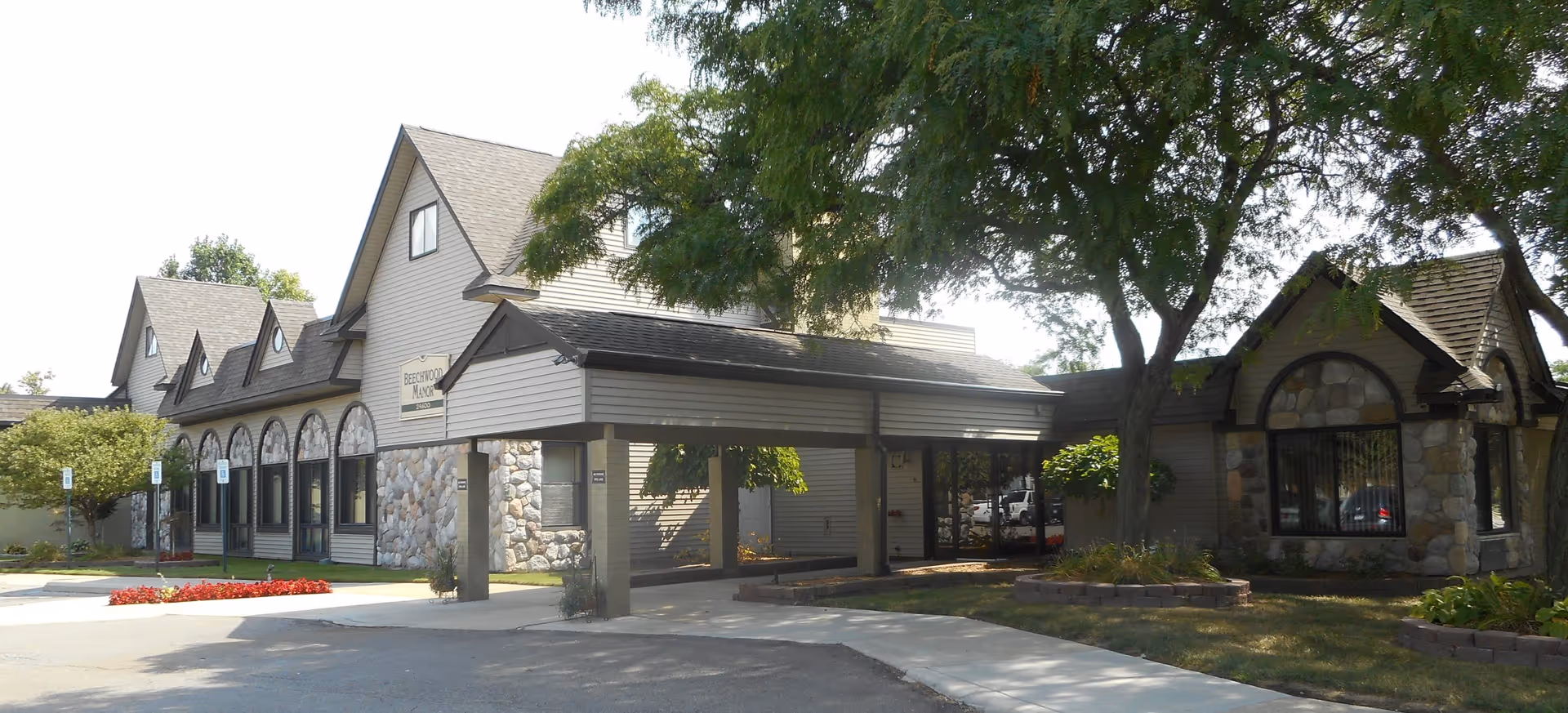 Exterior view of Beechwood Manor Inc, a building with stone and siding facade, multiple peaked roofs, large windows, and a covered entrance. There are trees and landscaped areas with flowers and shrubs around the building.
