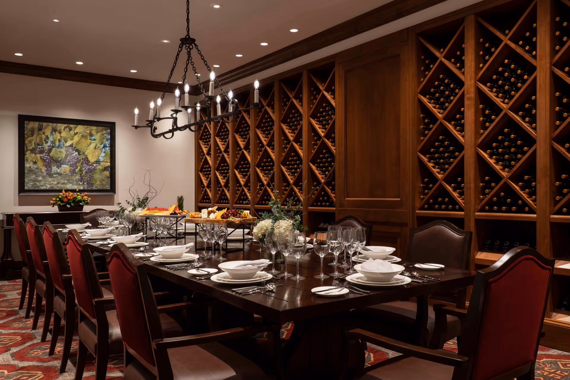A formal dining room with a long wooden table set with white plates, bowls, silverware, and wine glasses. The room features a large wooden wine rack filled with bottles along one wall, a chandelier hanging above the table, and a framed painting of grapes on the wall. There are red upholstered chairs around the table and a patterned carpet on the floor.
