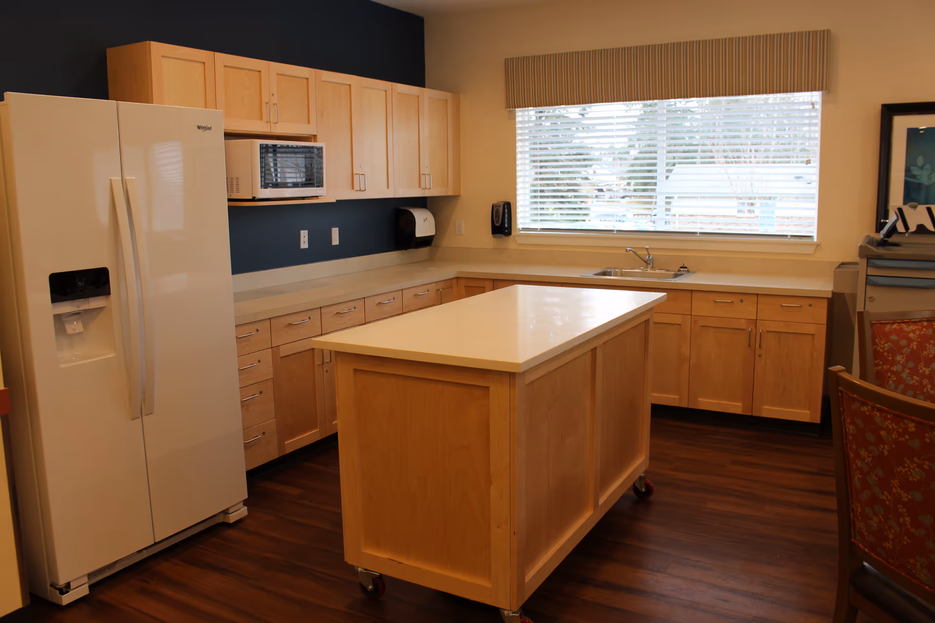 Bright communal kitchen with a central island, refrigerator, microwave, sink, and light wooden cabinets.