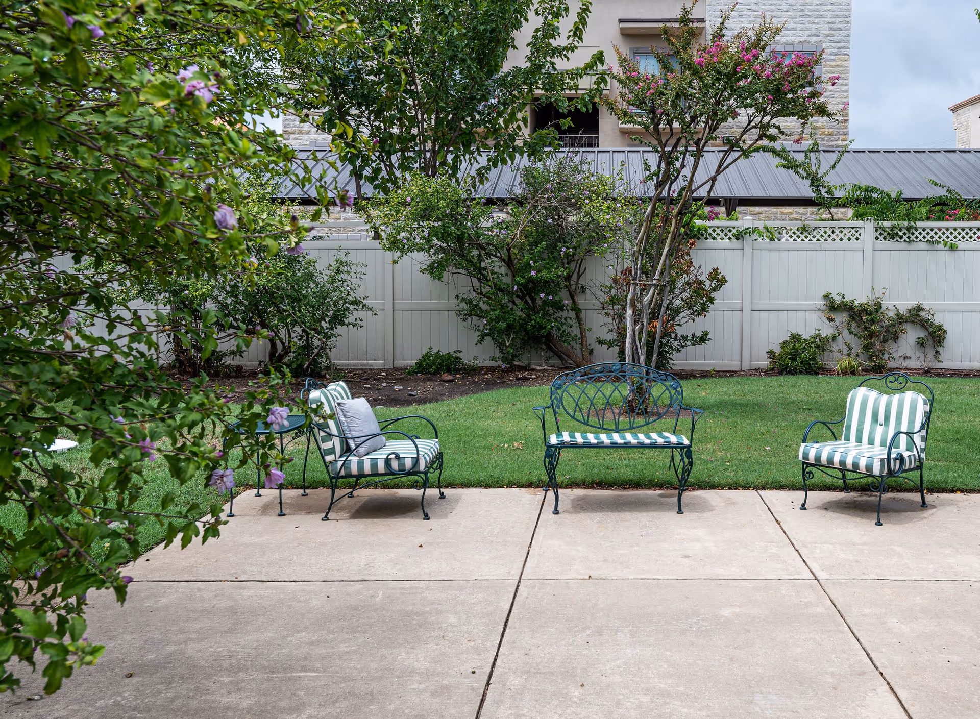 Outdoor patio area with three metal chairs and a bench with green and white striped cushions, surrounded by green grass, trees, and bushes, with a white fence and building in the background.