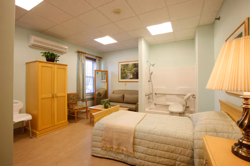 Furnished nursing facility bedroom with a bed, wardrobe, seating area and an accessible shower-bathtub.