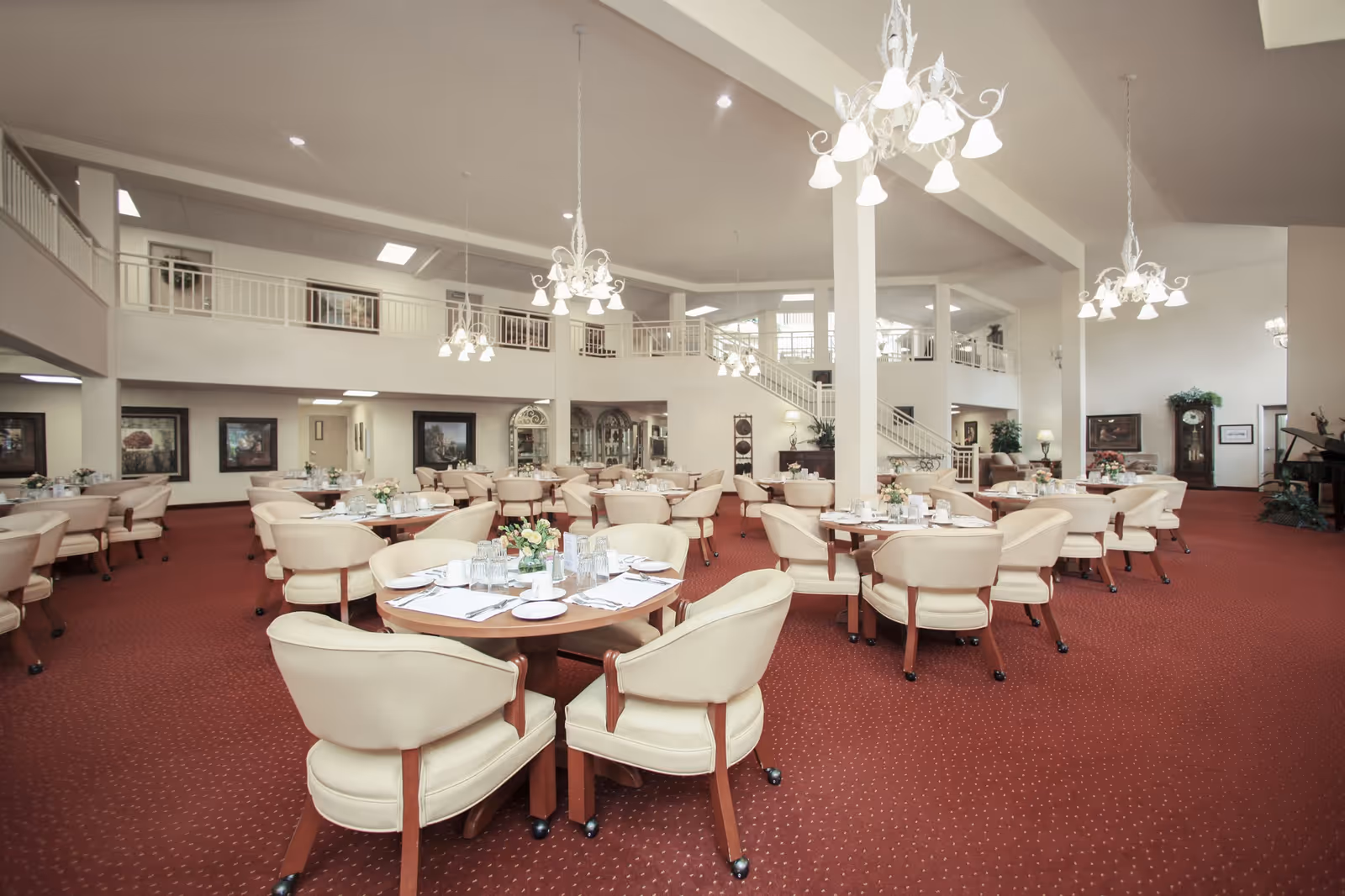 Spacious dining room with multiple round tables set with plates, glasses, and napkins. The room features cream-colored cushioned chairs, red carpet flooring, white walls, chandeliers hanging from the ceiling, and a staircase leading to an upper level with railings.
