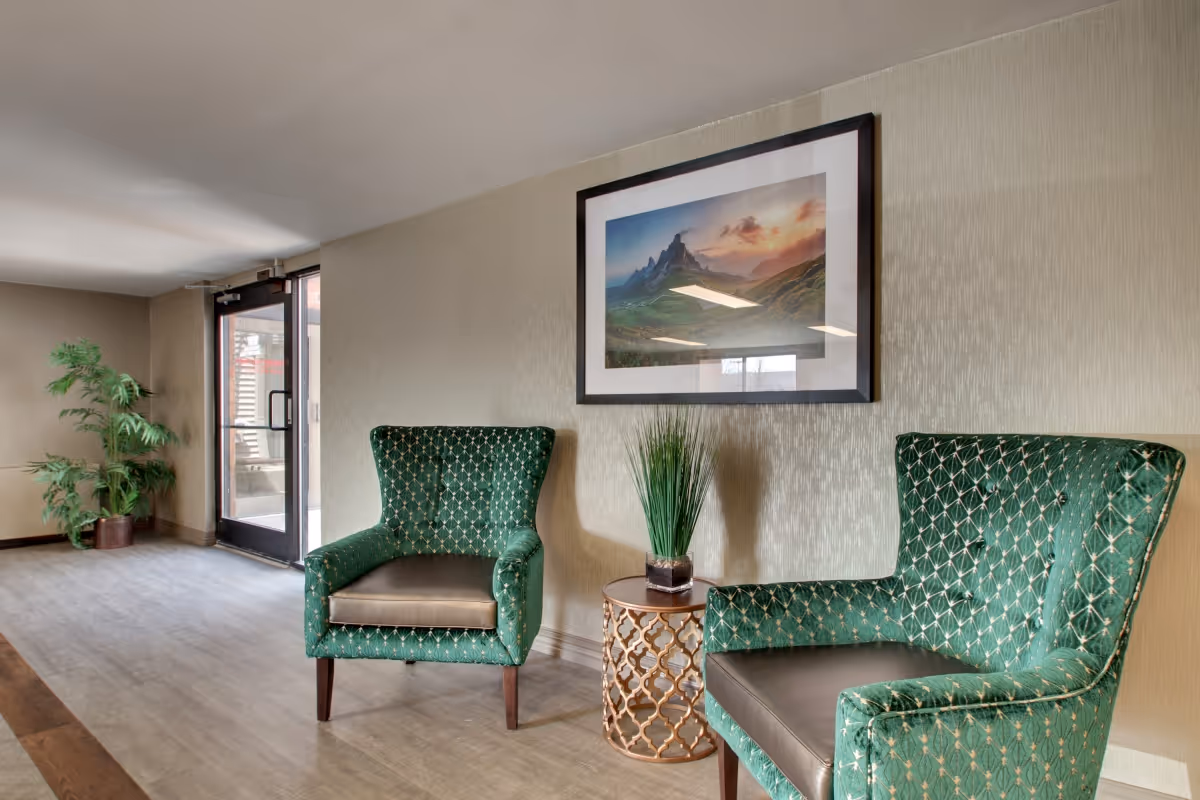A seating area in a senior living facility with two green upholstered armchairs with patterned fabric, a small round side table with a decorative plant, a framed landscape picture on the wall, and a glass door leading outside. There is also a potted plant in the corner.