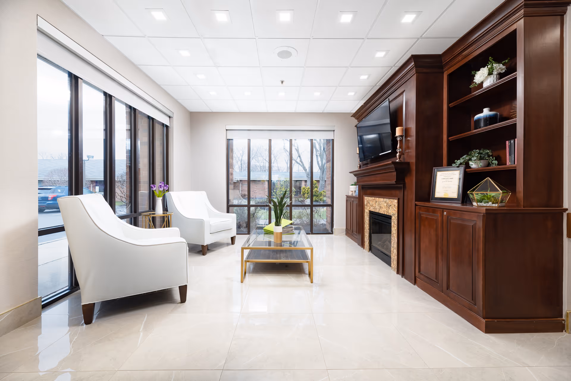 A bright and modern living room area with large floor-to-ceiling windows on two sides, two white armchairs, a glass coffee table with a plant, and a dark wood built-in shelving unit with a fireplace and a mounted flat-screen TV.