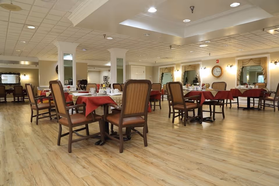 Spacious dining room with multiple tables set with red tablecloths and wooden chairs on a wood floor.