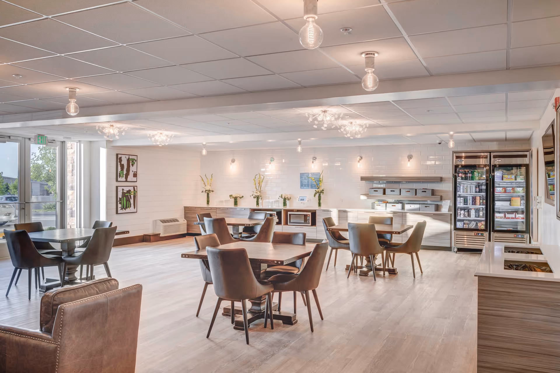 Bright communal dining area with several tables and chairs, a service counter, and refrigerated display cases.