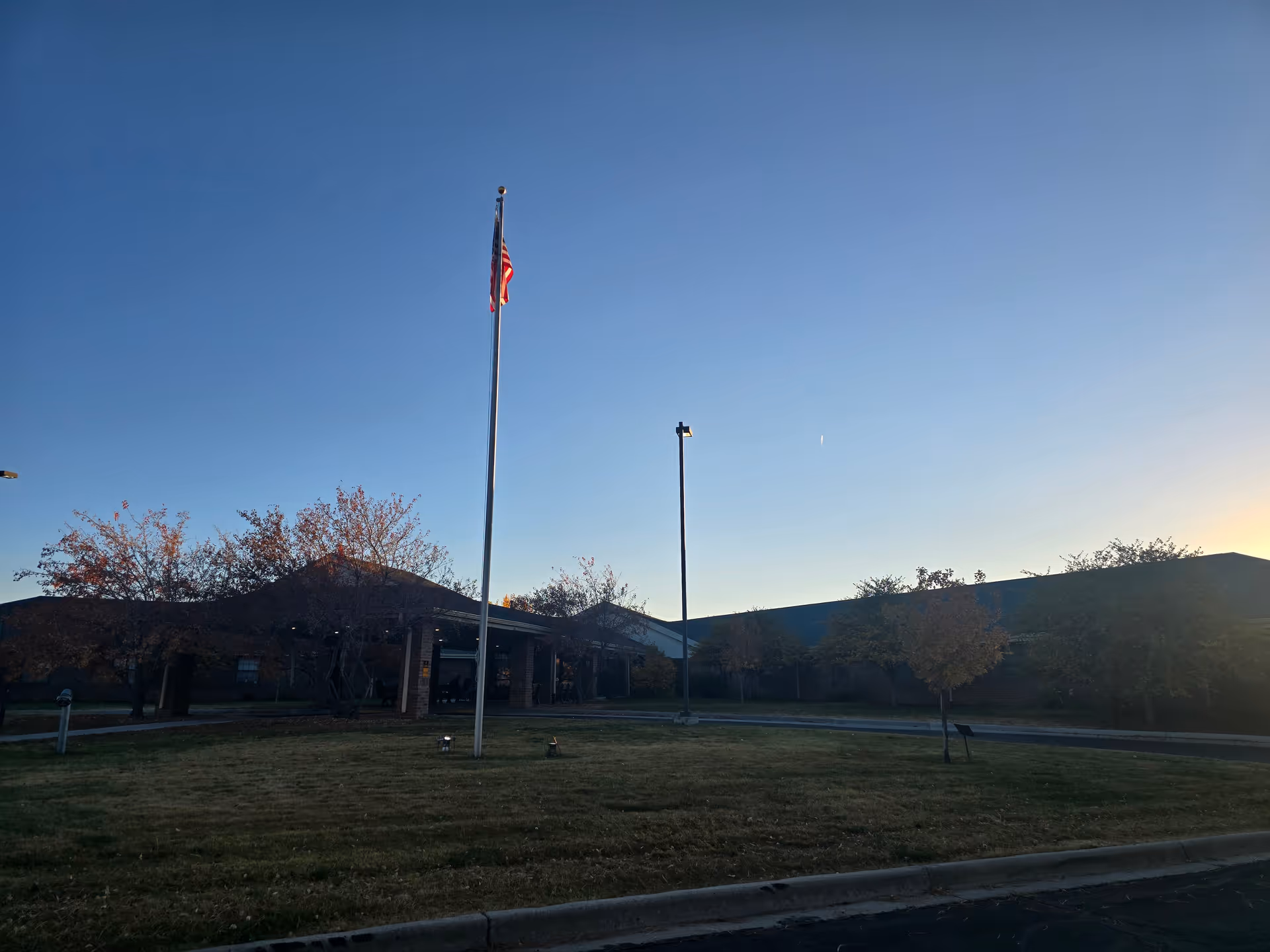 Exterior view of Brookside Inn facility during early evening with a clear sky, an American flag on a tall flagpole in the center, trees with autumn leaves, and a building with a covered entrance.