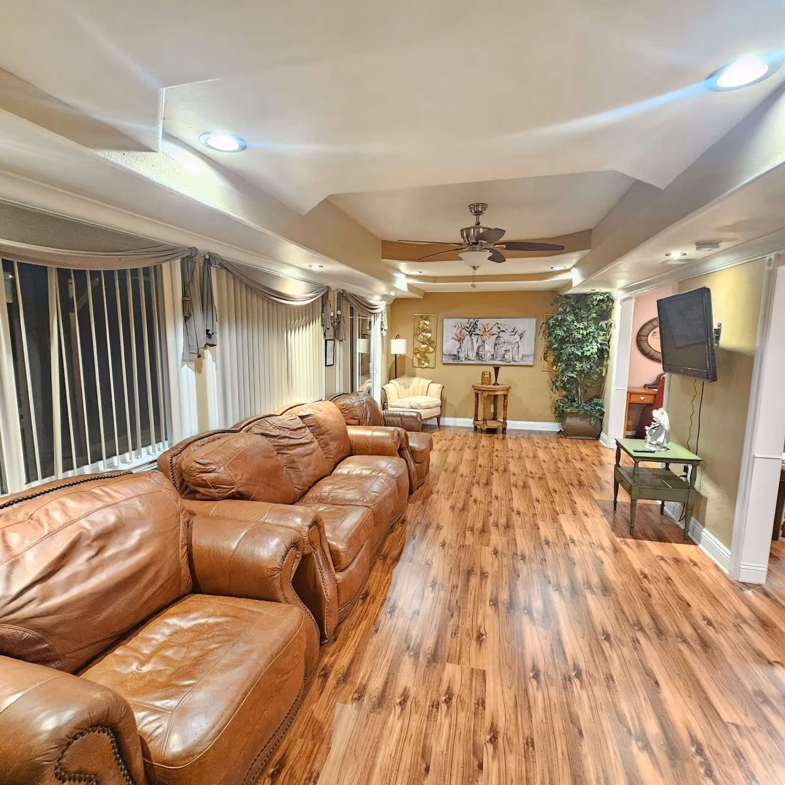 A spacious living room with wooden flooring, three brown leather sofas lined up along the left wall with vertical blinds on the windows behind them. The ceiling has recessed lighting and a ceiling fan. At the far end of the room, there is a small round wooden table with a lamp and decorative items, a beige armchair, a large potted plant, and a wall-mounted flat-screen TV on the right wall.
