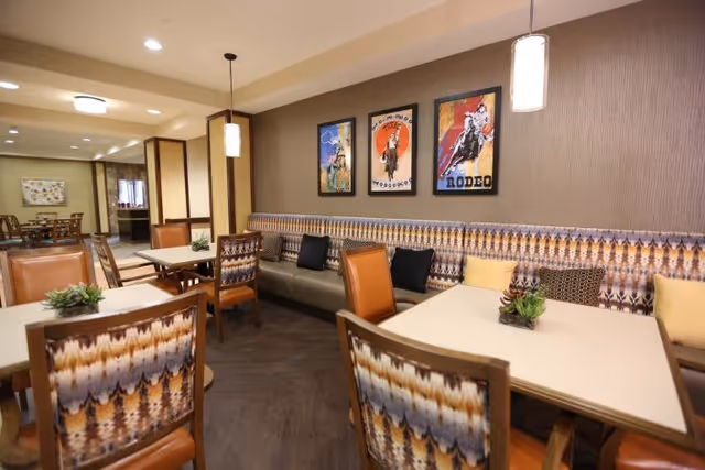 Interior view of a dining area with multiple tables and chairs. The chairs have patterned upholstery and some are leather. There is a long cushioned bench along the wall with decorative pillows. Three framed posters with rodeo themes hang on the wall above the bench. Pendant lights hang from the ceiling, illuminating the space.