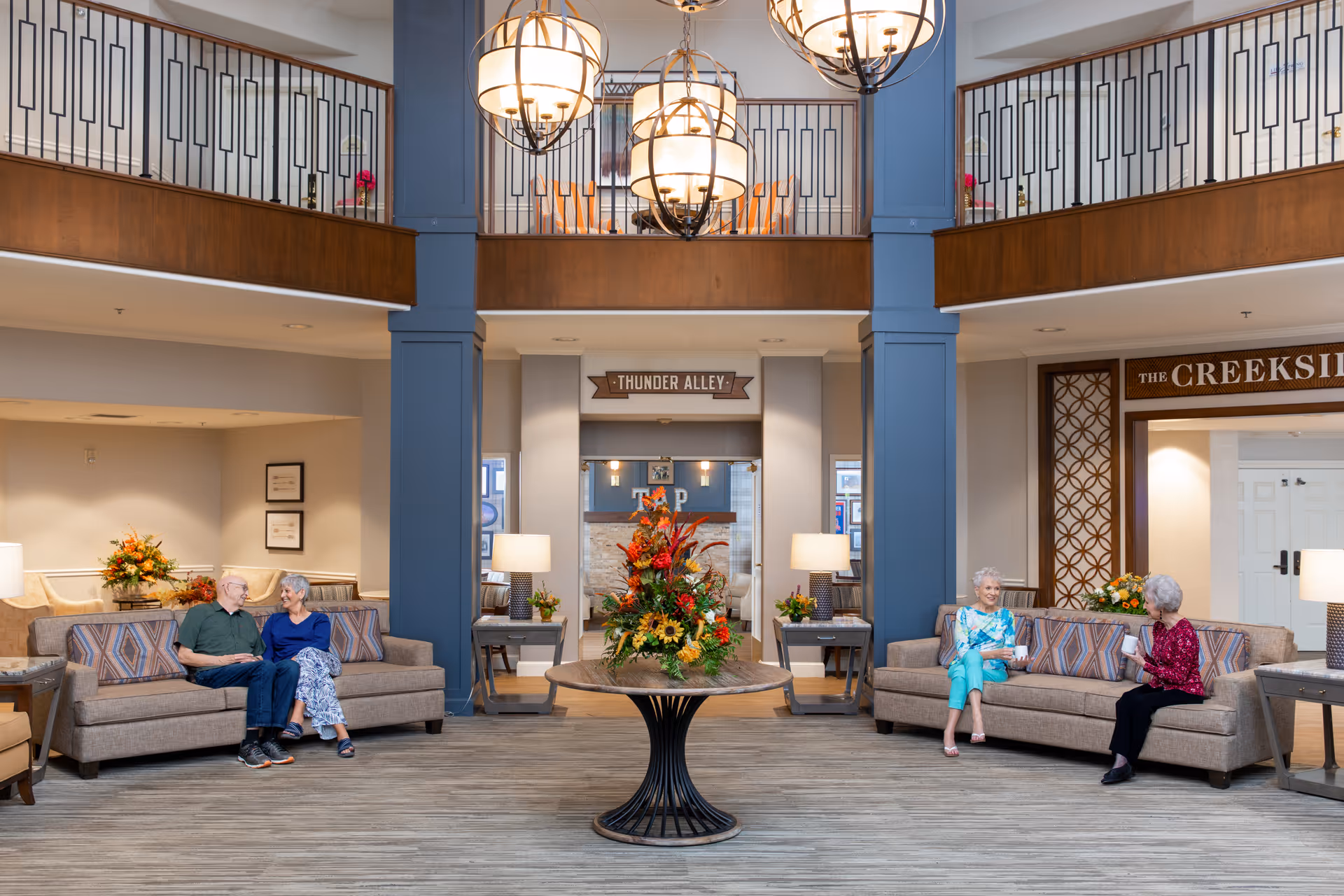 A spacious and well-lit senior living facility common area with two beige sofas facing each other. On the left sofa, an elderly man and woman sit closely, smiling and conversing. On the right sofa, two elderly women are seated, also engaged in conversation. In the center of the room is a round table with a large floral arrangement. The room features tall blue columns, a second-floor balcony with decorative railings, and three large hanging light fixtures. Signs reading 'THUNDER ALLEY' and 'THE CREEKSIDE' are visible in the background.