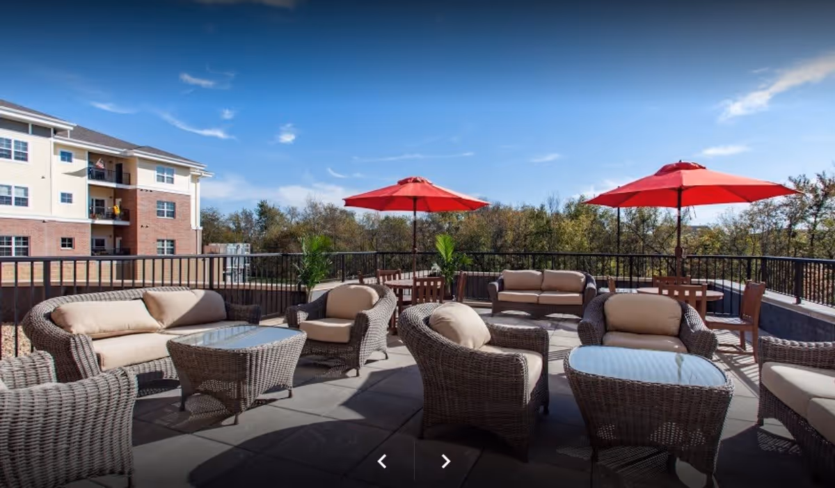 Outdoor patio area with wicker furniture including cushioned chairs and sofas, glass-top tables, and red umbrellas. The patio overlooks trees and a multi-story building under a clear blue sky.