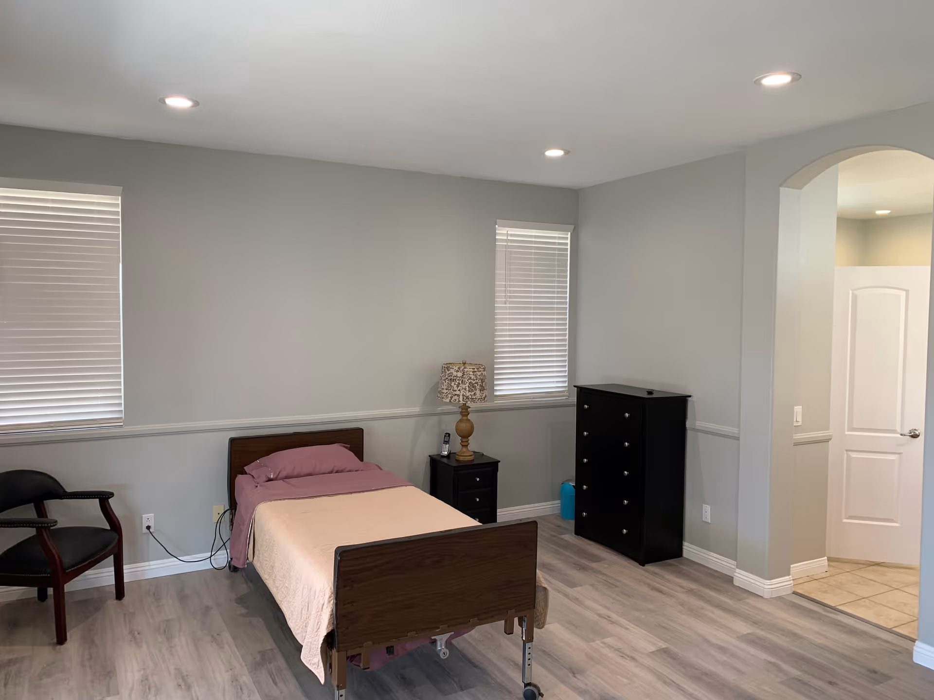 A simple bedroom with a single bed covered with a light pink blanket and a purple pillow. There is a wooden nightstand with a decorative lamp and a phone next to the bed. A black dresser with multiple drawers is against the wall. Two windows with closed blinds are on the wall behind the bed. A black chair is positioned near the left side of the bed. The floor is light wood, and the walls are painted light gray. There is an open doorway leading to another room with tiled flooring.
