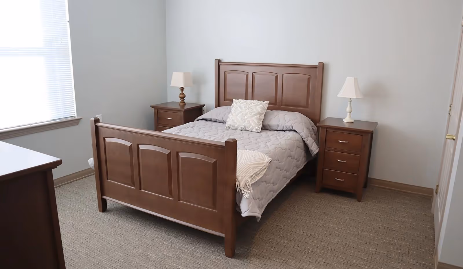 A simple bedroom with a wooden bed frame, a gray quilted bedspread, and a decorative pillow. There are two matching wooden nightstands on either side of the bed, each with a lamp. The room has light gray walls, a window with blinds, and beige carpeted flooring.