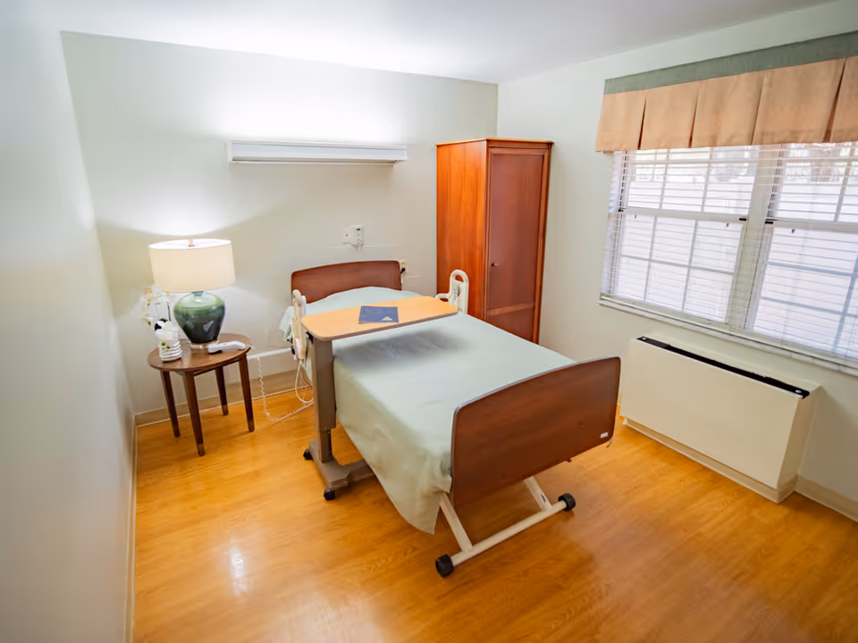 A small, neatly furnished bedroom in a senior living facility with a single bed, nightstand, lamp, wardrobe, and window blinds.