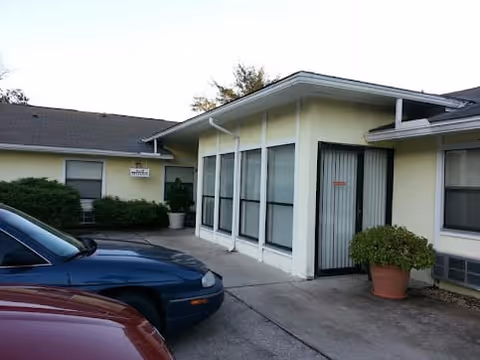 Exterior view of a single-story building with light yellow walls and multiple windows. There are two cars parked in front, a blue car and a red car. A potted plant is placed near a door with a metal security gate. Some bushes are visible near the building.