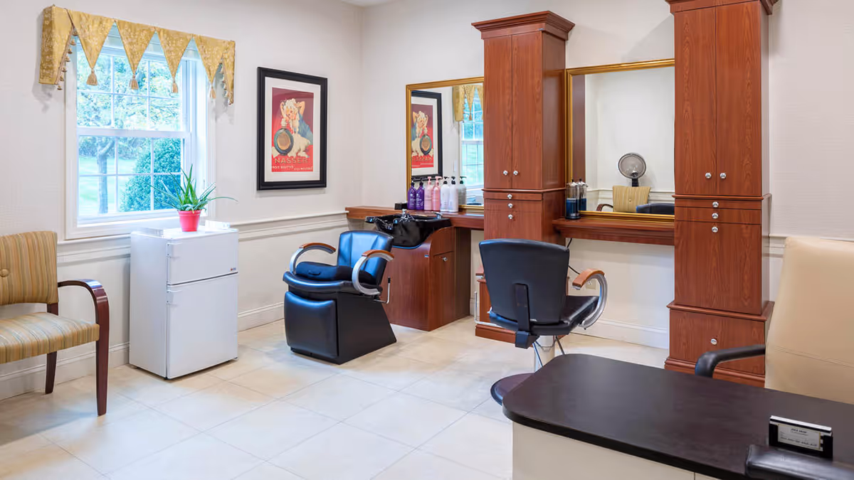 Interior of a salon area with two salon chairs facing a large mirror mounted on a wooden cabinet. There is a small white refrigerator with a potted plant on top near a window with a yellow valance. Two chairs, one striped and one beige, are positioned around the room. Various hair care products are placed on the counter near the mirror.