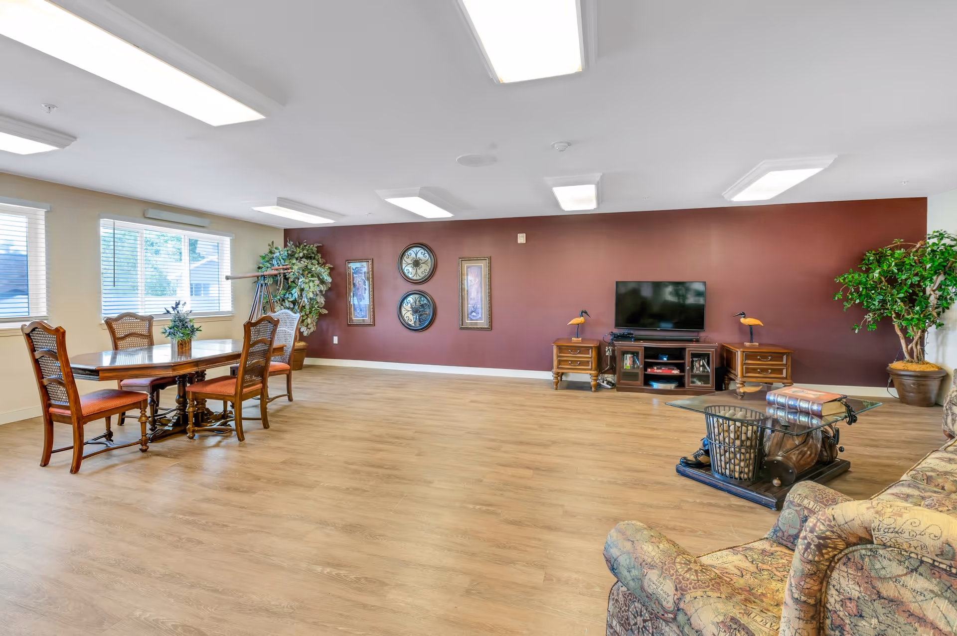 A spacious living room with wooden flooring, a maroon accent wall, and beige walls with windows. The room features a wooden dining table with four chairs, a TV on a wooden stand flanked by two small wooden cabinets with decorative bird lamps, a glass coffee table with books on it, and a patterned armchair. There are also two large potted plants and framed artwork on the accent wall.