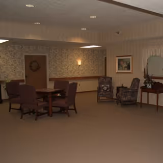 A common area in a senior living facility with a table surrounded by six chairs on the left and two upholstered armchairs with a small table between them on the right. The walls have floral wallpaper and there is a framed picture and a mirror above a wooden console table. The room is carpeted and softly lit with ceiling lights and a wall sconce.