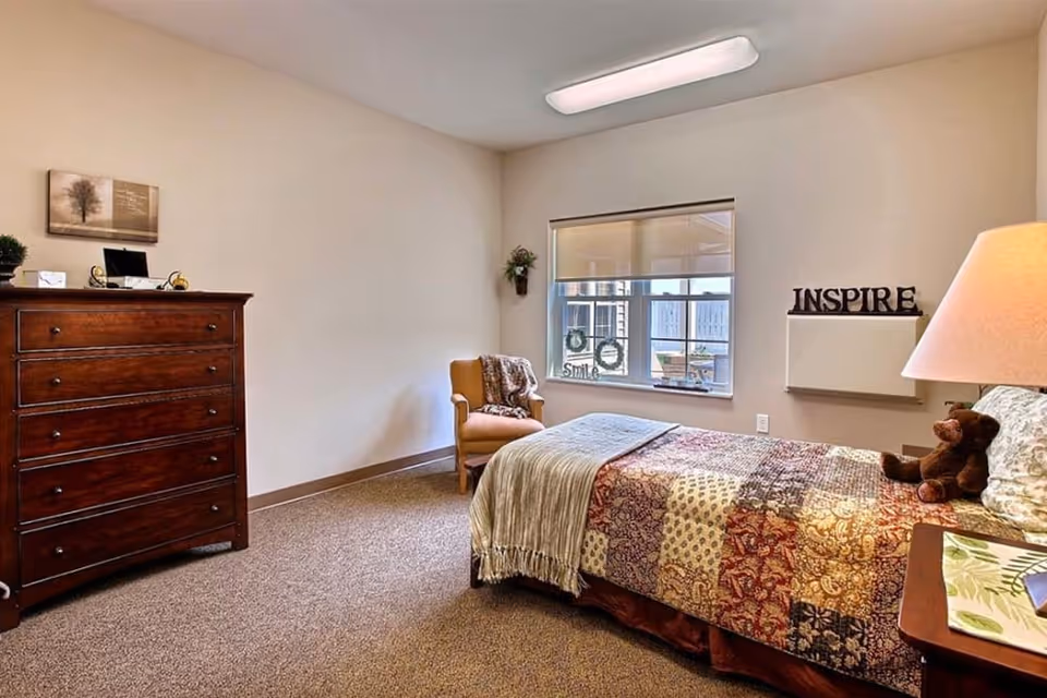 A cozy senior living bedroom with a single bed covered in a colorful patchwork quilt, a wooden nightstand with a lamp and a teddy bear, a wooden chest of drawers, a comfortable armchair near a window with a view outside, and decorative items including a wall art, a small plant, and the word 'INSPIRE' displayed on a shelf.