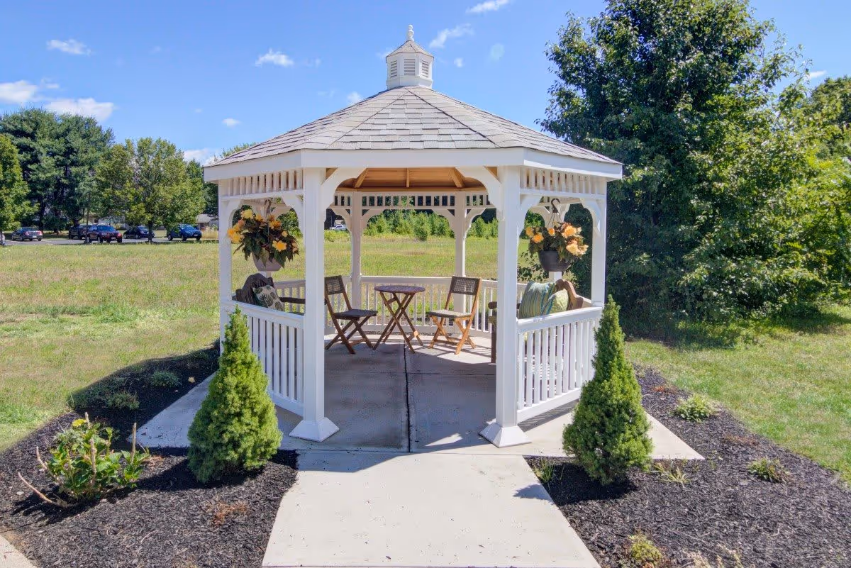 A white wooden gazebo with a shingled roof situated outdoors on a concrete pad surrounded by small landscaped bushes and mulch. Inside the gazebo are wooden chairs and a small table, with hanging flower pots on the posts. The background shows a grassy field, trees, and a few parked cars under a blue sky with some clouds.