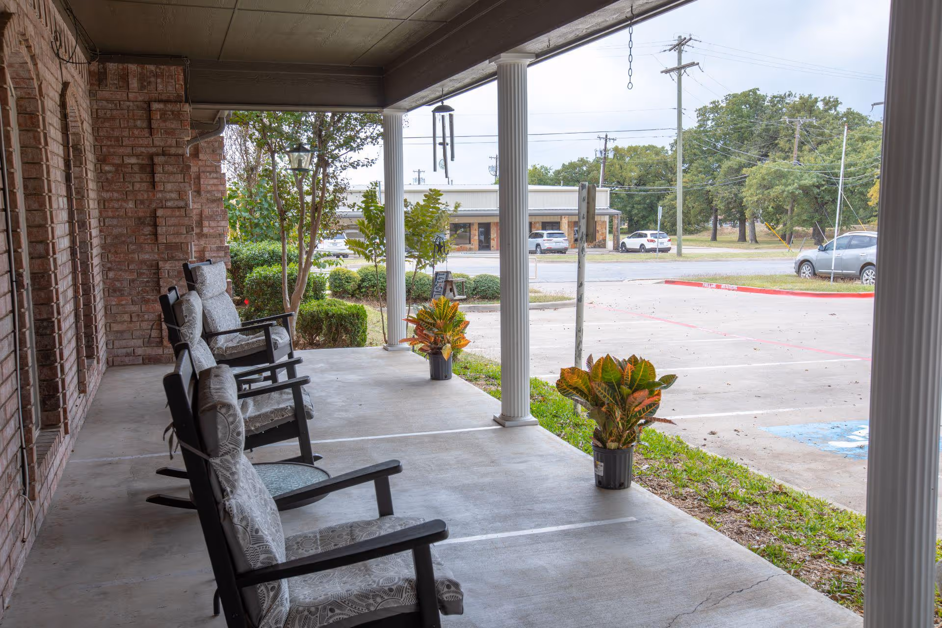 Covered outdoor porch area with cushioned rocking chairs lined up facing outward. There are potted plants placed near white columns supporting the roof. In the background, a parking lot with several cars and a small commercial building across the street are visible. Trees and greenery surround the area.