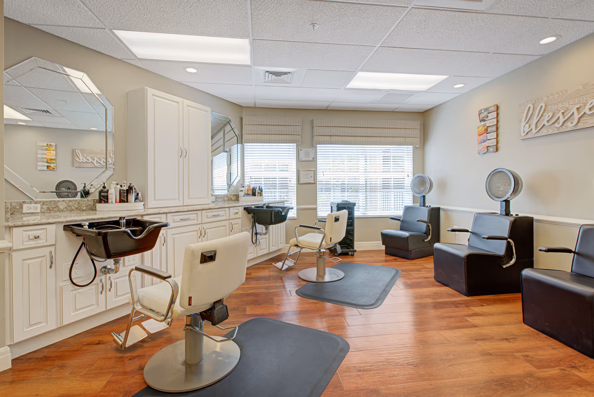 Interior view of a hair salon area in a senior living facility with two white salon chairs in front of black hair washing sinks, large mirrors, white cabinetry, and three black waiting chairs along the wall. The room has wooden flooring, large windows with blinds, and wall decorations including a sign that says 'blessed'.
