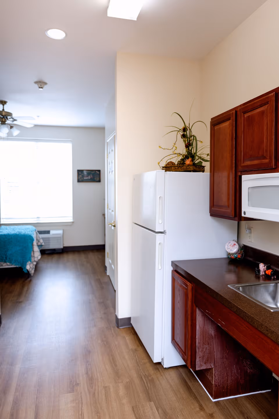 Small kitchenette with a white refrigerator, dark wood cabinets and sink opening into a studio bedroom area with a bed by a window.