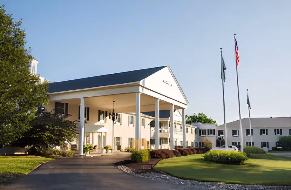 Front exterior view of a large white building with a covered entrance supported by tall white columns, a driveway leading up to it, well-maintained green lawns, shrubs, and three flagpoles with flags flying against a clear blue sky.