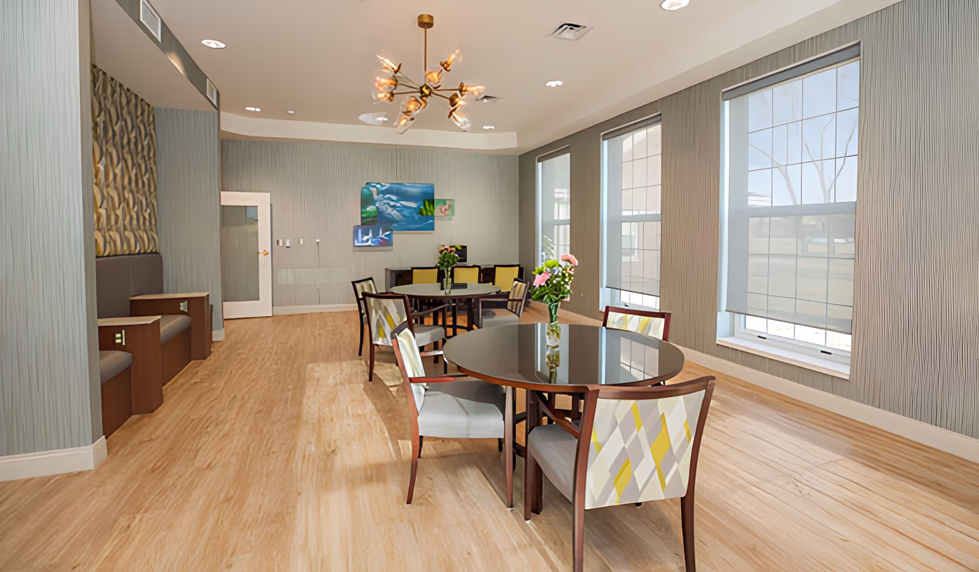 Bright common dining area with round tables, patterned chairs, large windows, and modern lighting.