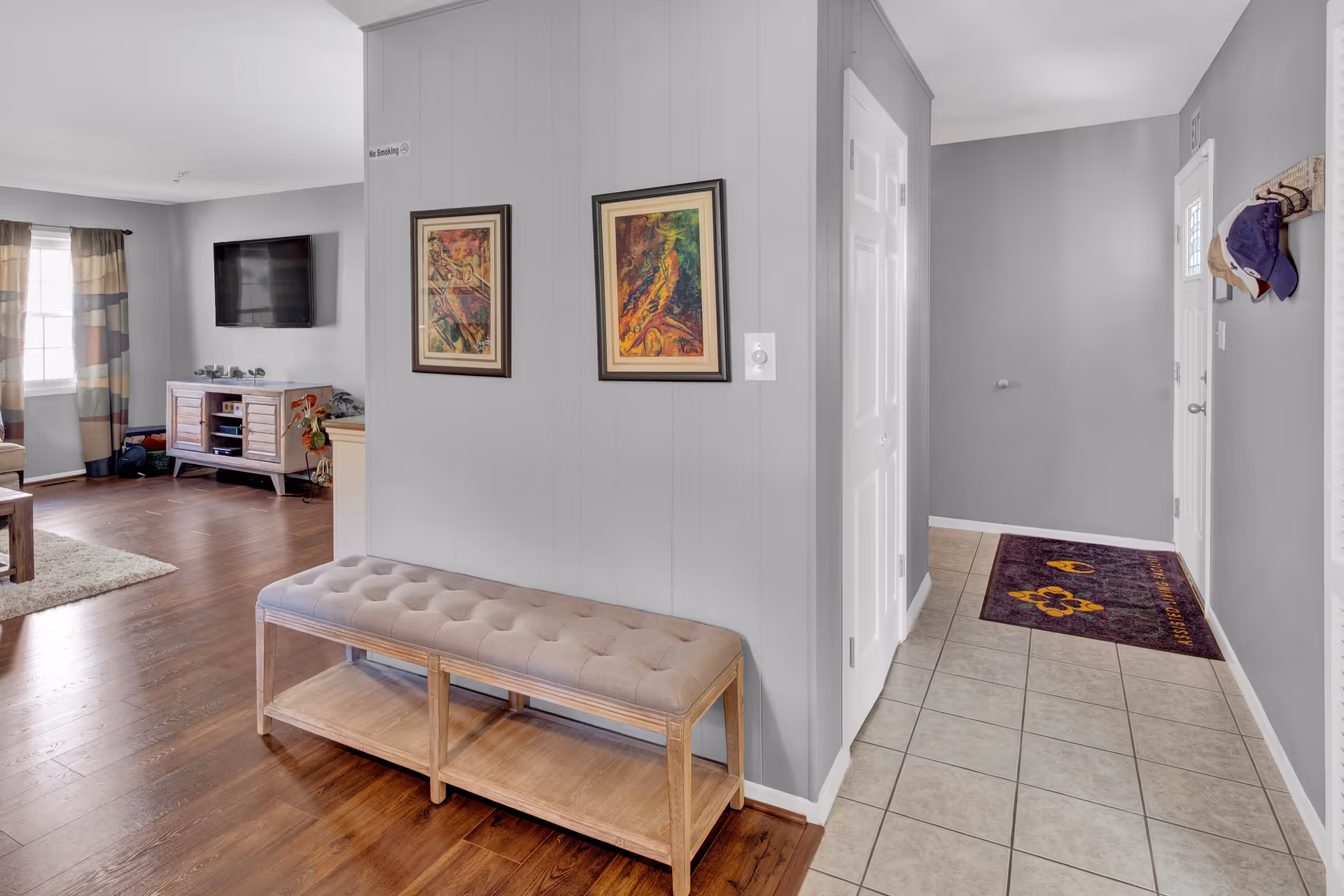 Interior view of a senior living facility showing a hallway with tiled floor leading to a door with a purple mat. The walls are painted gray with two framed colorful paintings and a 'No Smoking' sign. A cushioned wooden bench is placed against the wall. To the left, there is a living room area with wooden flooring, a TV mounted on the wall, a wooden TV stand, and a window with patterned curtains.