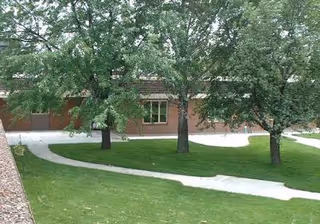Outdoor view of a green lawn with several mature trees and a winding concrete pathway in front of a brick building with windows and garage doors.