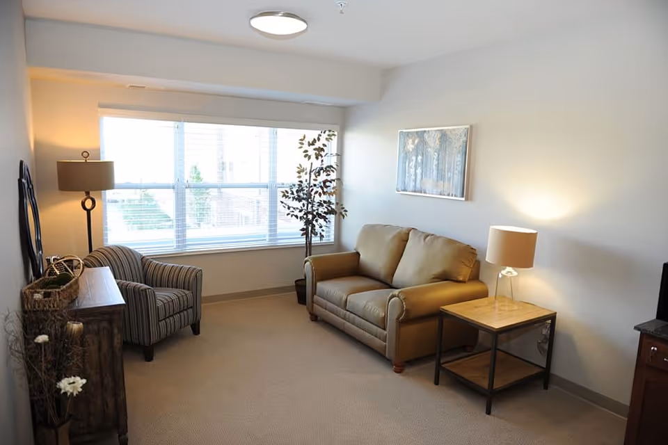 A cozy living room with a beige couch, a striped armchair, a wooden side table with a lamp, a floor lamp, a potted plant near a large window with blinds, and a framed artwork on the wall.