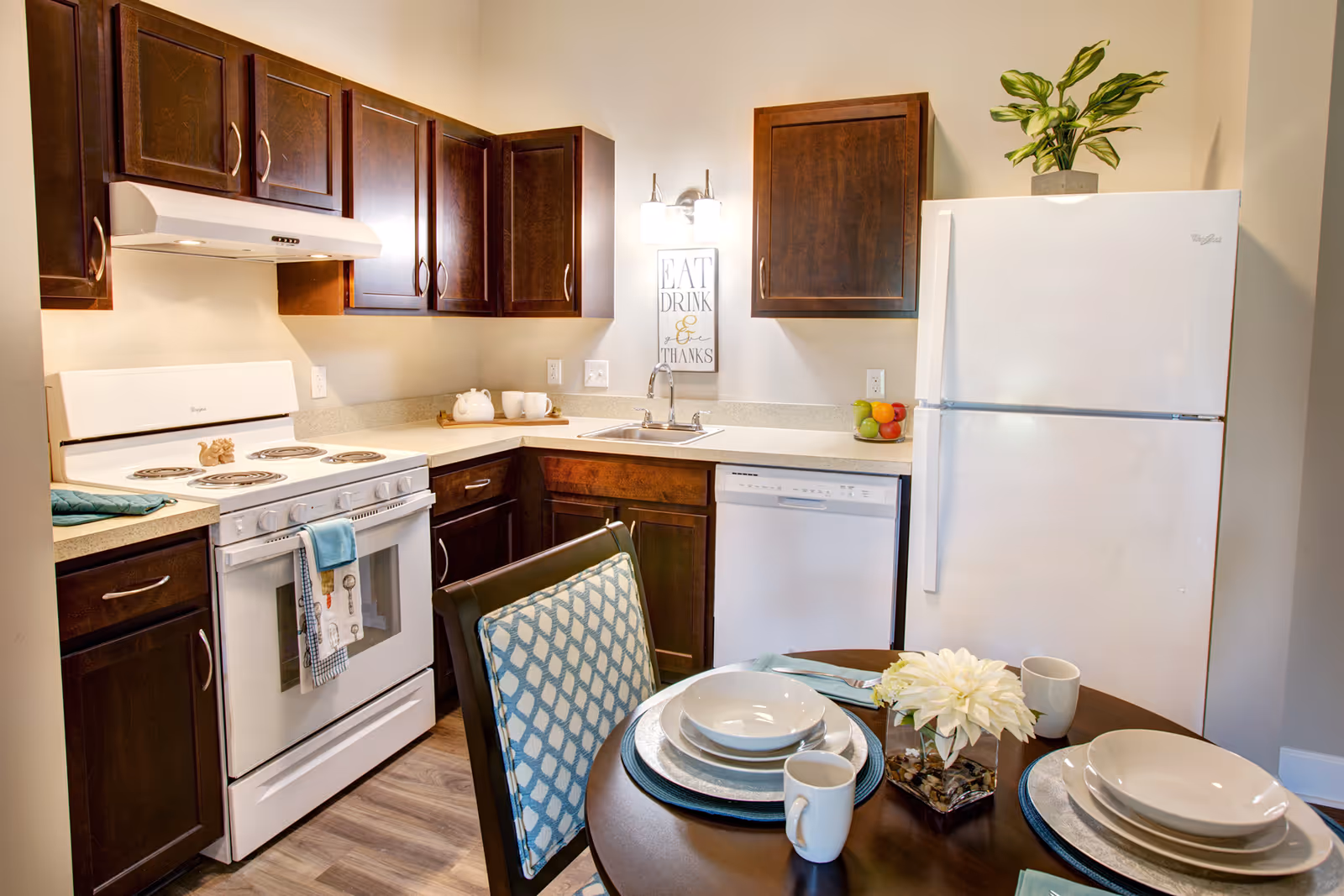 A bright kitchen with dark wood cabinets, a white electric stove with oven, a white refrigerator with a green plant on top, a dishwasher, and a sink under a wall light fixture. A small round dining table is set with white dishes, mugs, and a vase with white flowers. A decorative sign on the wall reads 'EAT DRINK & THANKS.'