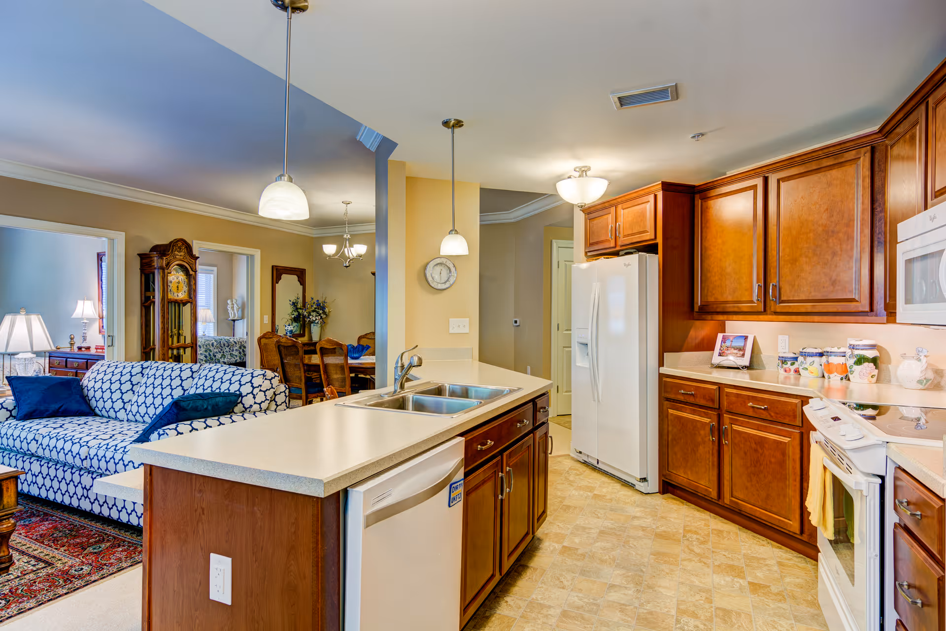 A bright and spacious kitchen with wooden cabinets, a white refrigerator, a white stove, and a double sink on a central island. The kitchen opens into a living area with a patterned blue and white sofa and a dining room with a wooden dining table and chairs. The space is well-lit with pendant lights and ceiling fixtures.