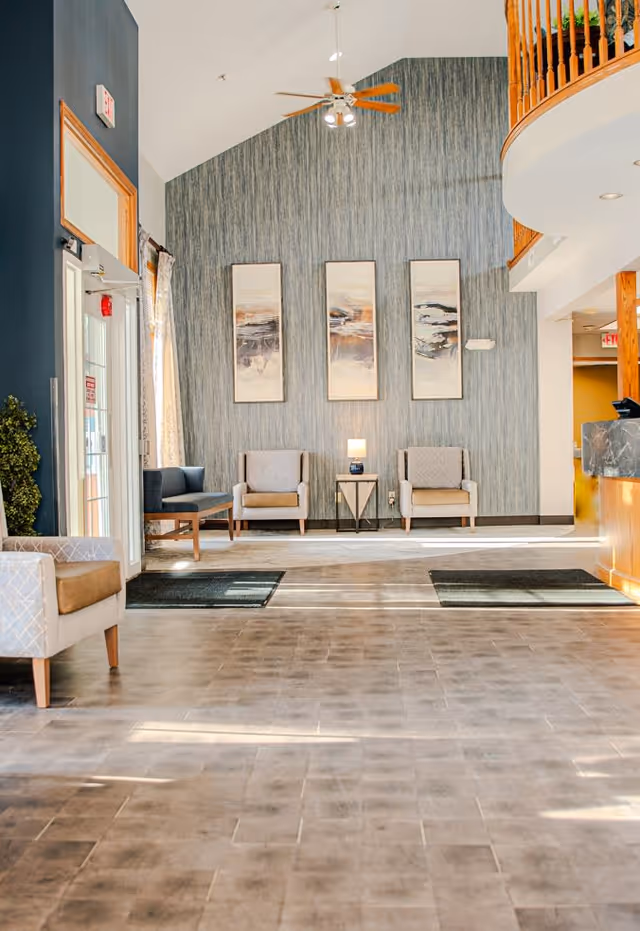 A bright and spacious lobby area with high ceilings, featuring a seating arrangement of armchairs and a small table with a lamp. The back wall has three vertical abstract paintings, and there is a ceiling fan above. The floor is tiled, and there are entrance mats near the door. A curved wooden railing is visible on the upper right side.