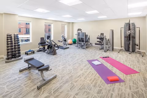 A well-lit exercise room with weight benches, machines, a rack of dumbbells, stability balls and two yoga mats on a carpeted floor.