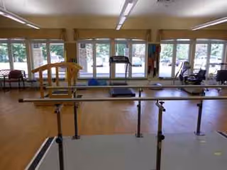 Empty therapy gym with parallel bars, treadmills, mats, and large windows in a senior living facility.