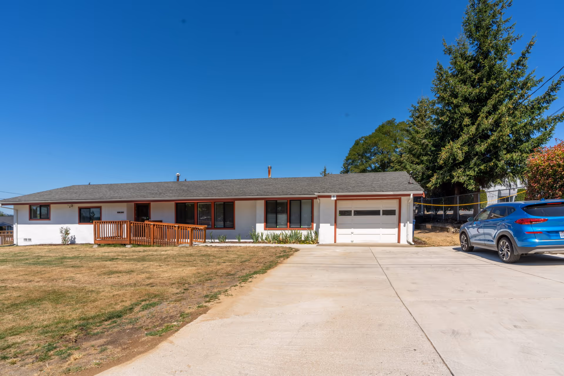 Single-story white building with a gray roof and a wooden ramp leading to the entrance, a driveway with a blue SUV parked on the right, and a large tree in the background under a clear blue sky.