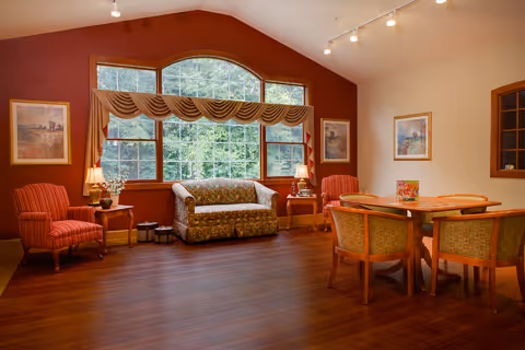 A cozy common area with wooden flooring featuring a large window with a decorative valance. The room contains a floral-patterned sofa, two red striped armchairs, two wooden side tables with lamps, and a wooden dining table with four green cushioned chairs. The walls are decorated with framed artwork.