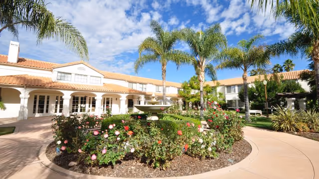Outdoor courtyard area of a senior living facility with a circular flower bed filled with blooming roses and a central fountain. The courtyard is surrounded by a two-story building with white walls and terracotta roof tiles. Several tall palm trees are scattered around the area under a partly cloudy blue sky.