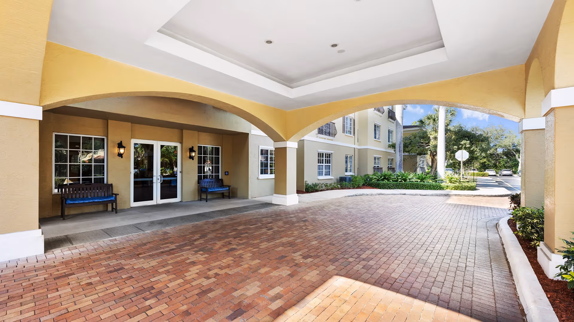 Covered entrance area of a senior living facility with brick-paved driveway, beige walls, two benches with blue cushions, double glass doors, and windows. The building exterior is light yellow with white trim, and there are plants and trees visible in the background under a clear sky.