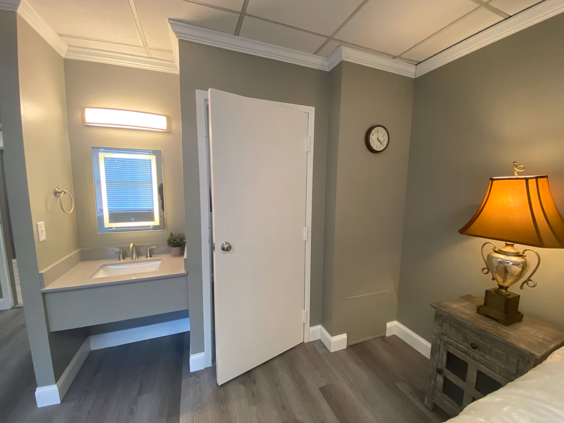 Interior view of a senior living facility room showing a small sink area with a mirror and light above it on the left, a partially open white door in the center, and a bedside table with a lamp on the right. The walls are painted gray and the floor has wood-style flooring.