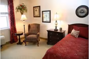 A cozy bedroom featuring a bed with a red patterned bedspread and a decorative pillow, a wooden nightstand with two lamps and small decorative items, an upholstered armchair, a small round side table, framed artwork on the wall, a window with red curtains, and a wall clock.