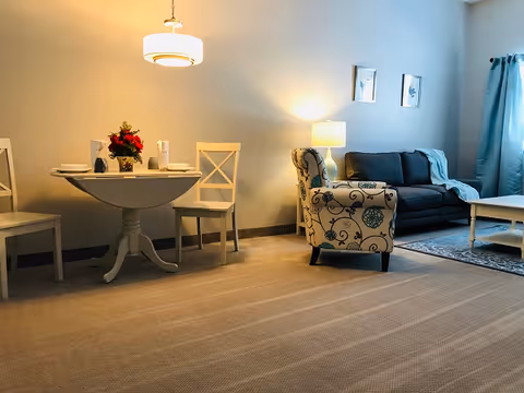 A cozy living area featuring a small round dining table set for two with white chairs and a floral centerpiece, a patterned armchair, a dark blue sofa with cushions, a white coffee table on a rug, and a lamp on a side table against a light-colored wall with two framed pictures.
