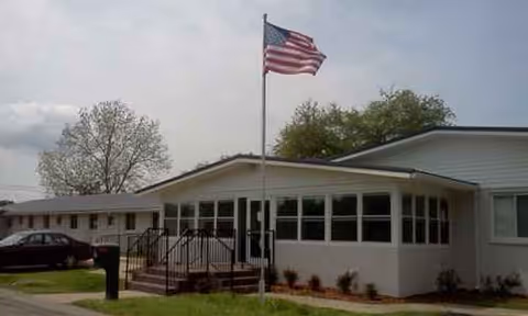 Single-story care facility front with flagpole, steps, and windows facing a green lawn.